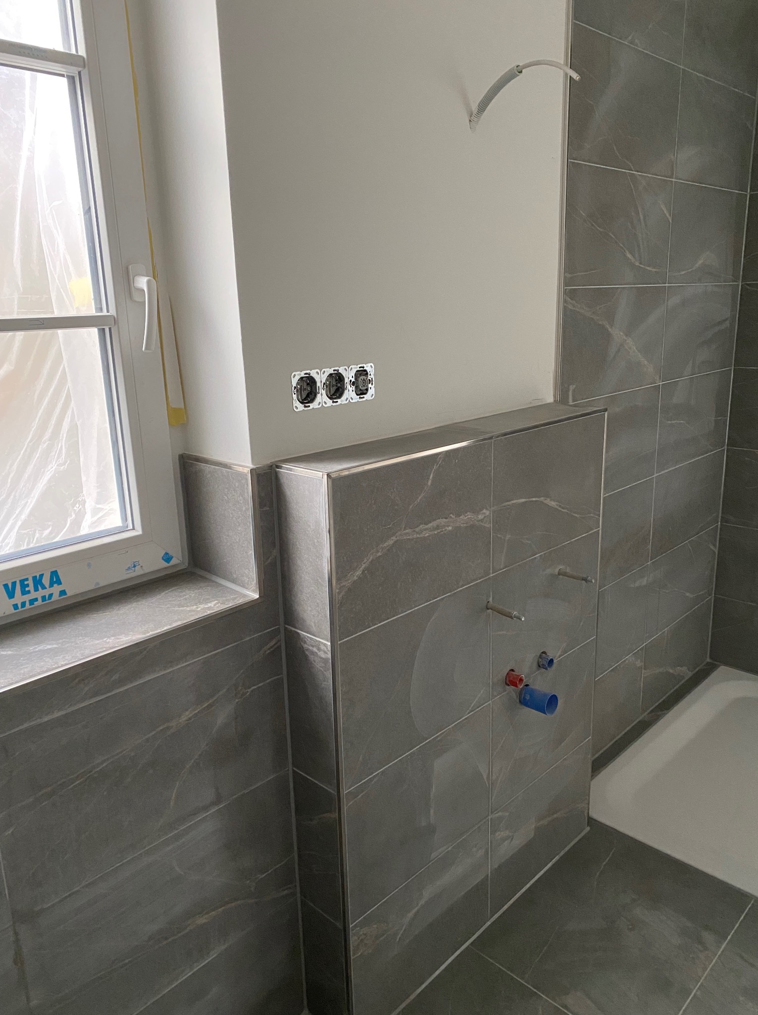 Wykończona łazienka z szarymi płytkami imitującymi marmur, widoczne przyłącza wodne i gniazdka elektryczne. Okno z folią ochronną w rogu.