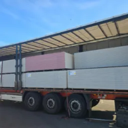 Ciężarówka z otwartą naczepą załadowana paletami płyt karton-gips, widoczne zabezpieczenia ładunku i błękitne niebo w tle. Część płyt jest w kolorze różowym.