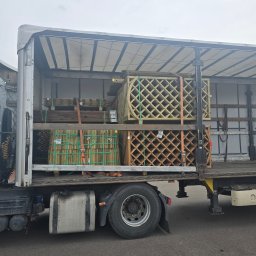 RS SPÓŁKA Z OGRANICZONĄ ODPOWIEDZIALNOŚCIĄ - Ciężarówka z otwartą naczepą, w której przewożone są drewniane elementy ogrodowe, zabezpieczone pasami. Widoczne koło i fragment kabiny.