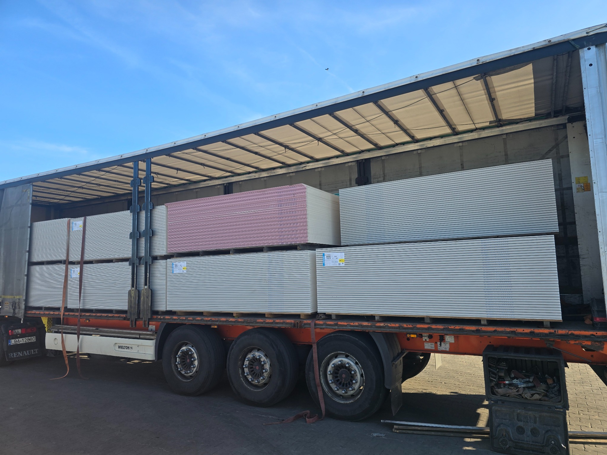 Ciężarówka z otwartą naczepą załadowana paletami płyt karton-gips, widoczne zabezpieczenia ładunku i błękitne niebo w tle. Część płyt jest w kolorze różowym.