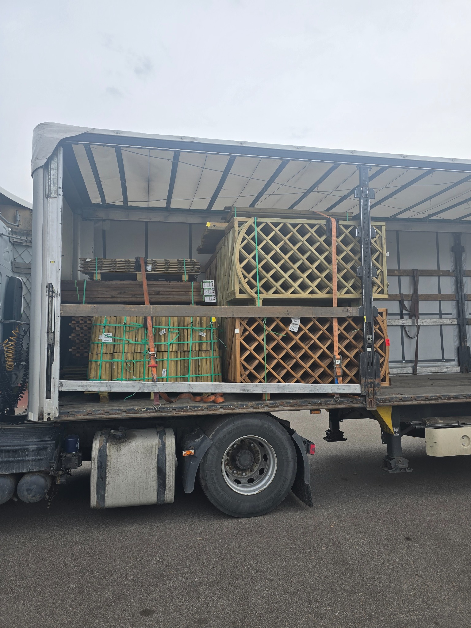 Ciężarówka z otwartą naczepą, w której przewożone są drewniane elementy ogrodowe, zabezpieczone pasami. Widoczne koło i fragment kabiny.