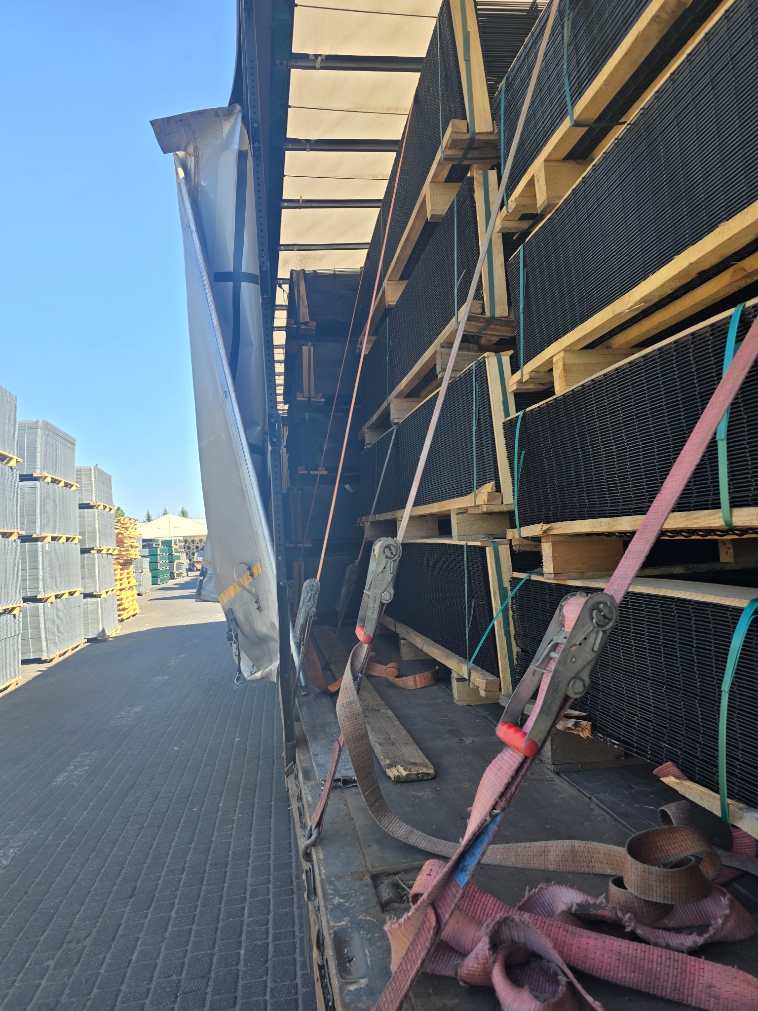 Ciężarówka załadowana paletami z czarnymi panelami siatkowymi, zabezpieczonymi pasami transportowymi. Na zewnątrz widoczne kolejne palety.