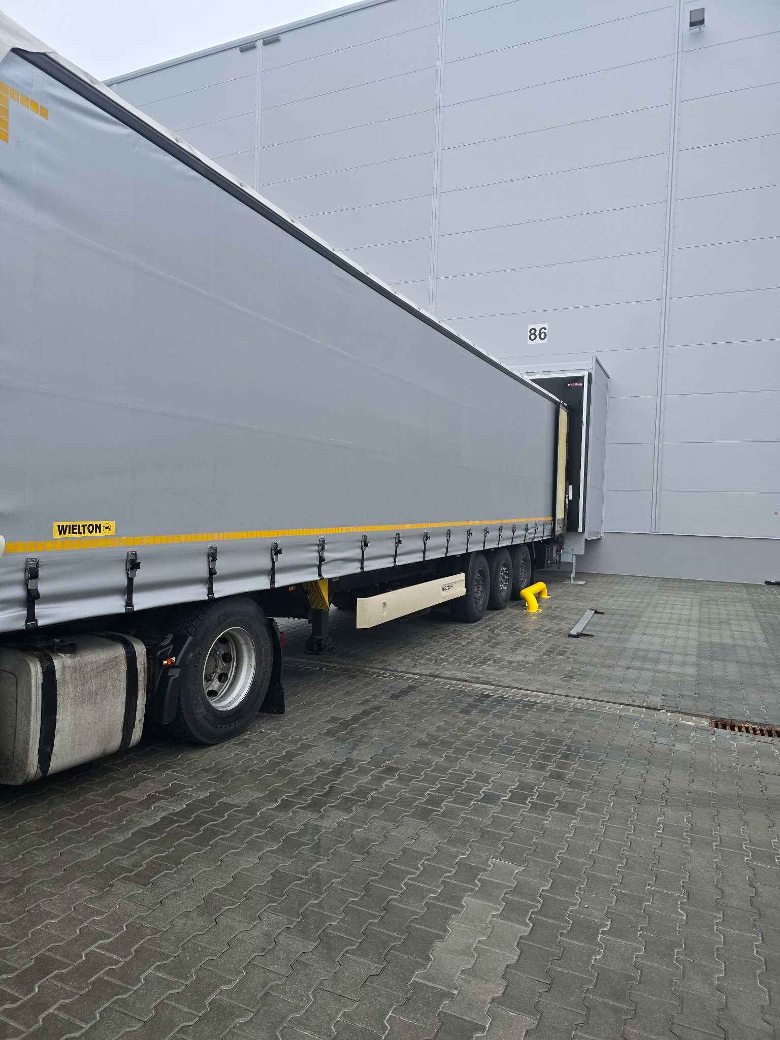 Ciężarówka marki Wielton przy rampie magazynowej nr 86 w Radomiu, gotowa do załadunku lub rozładunku. Widoczna kostka brukowa i fragment budynku magazynowego.