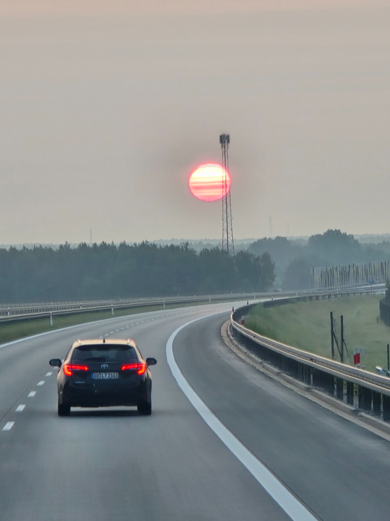 Samochód na autostradzie, w tle wschodzące słońce i wieża telekomunikacyjna. Widok z perspektywy kierowcy, światła tylne pojazdu włączone.