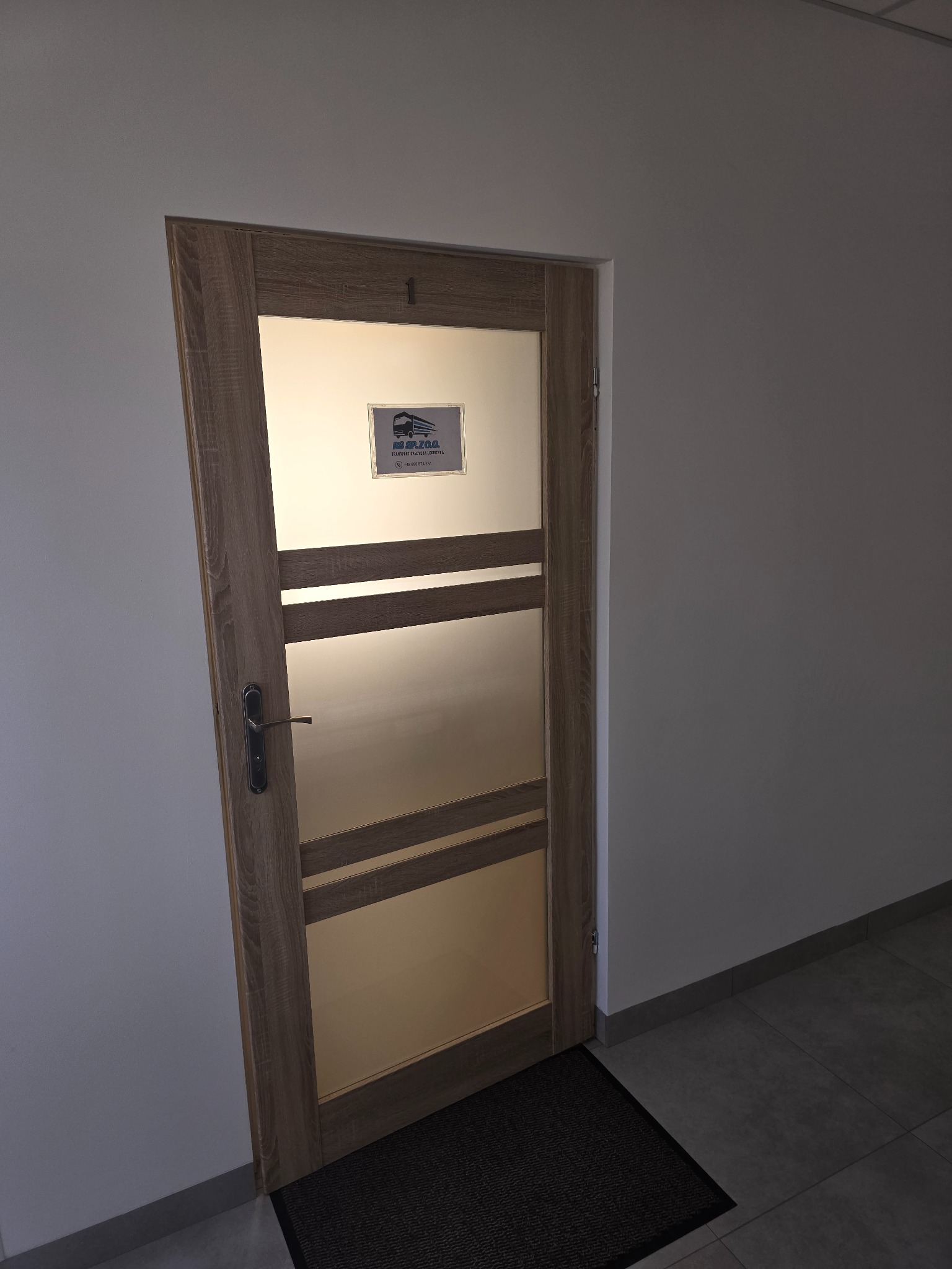 Drzwi wewnętrzne z numerem '1' i szybą z logo firmy transportowej. Czarny wycieraczka przed drzwiami. Widok na fragment korytarza z szarymi płytkami.