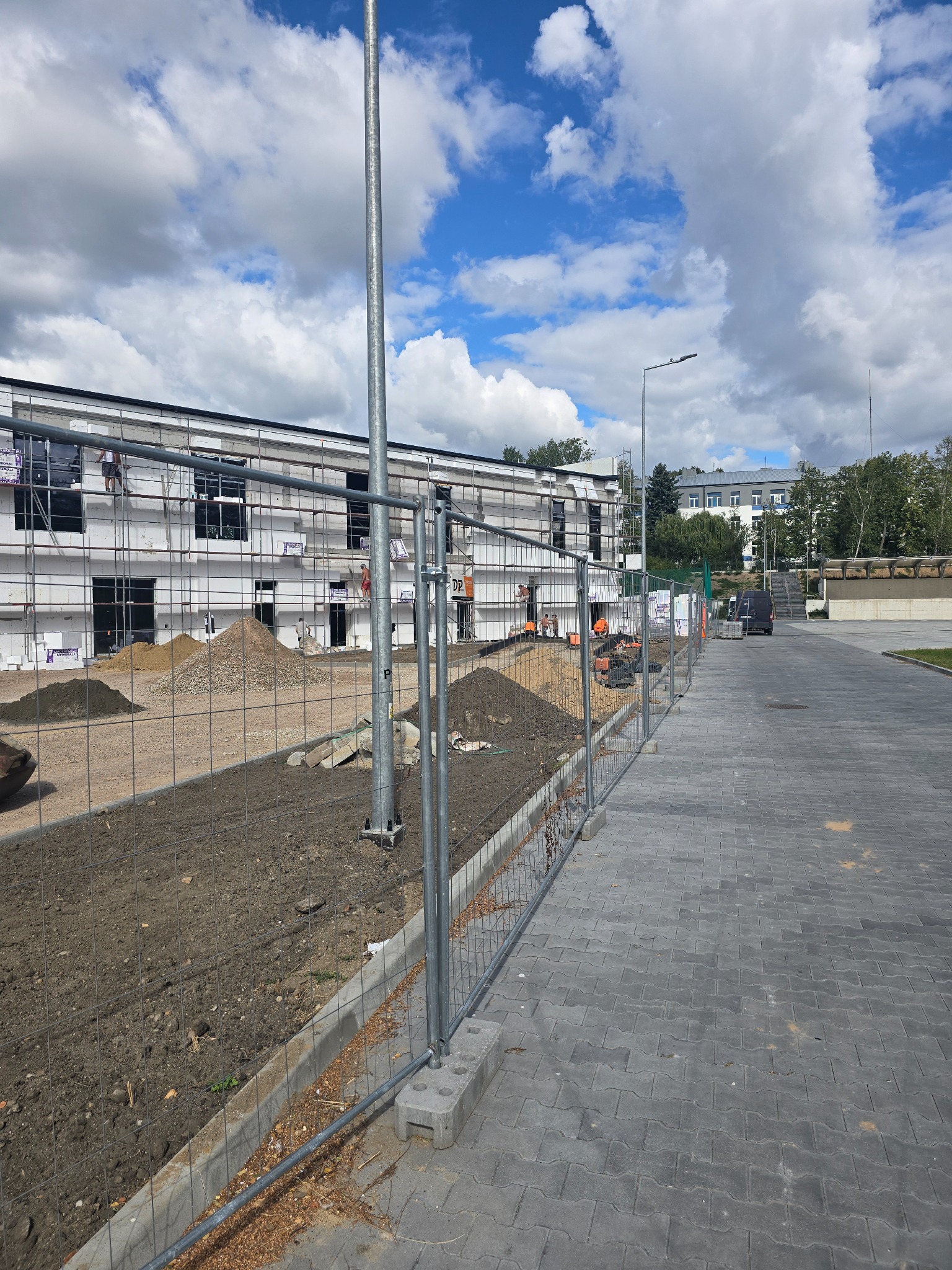 Budynek w trakcie ocieplania elewacji, widoczne rusztowania i pracownicy. Na pierwszym planie ogrodzenie budowy, hałda ziemi i kostka brukowa.