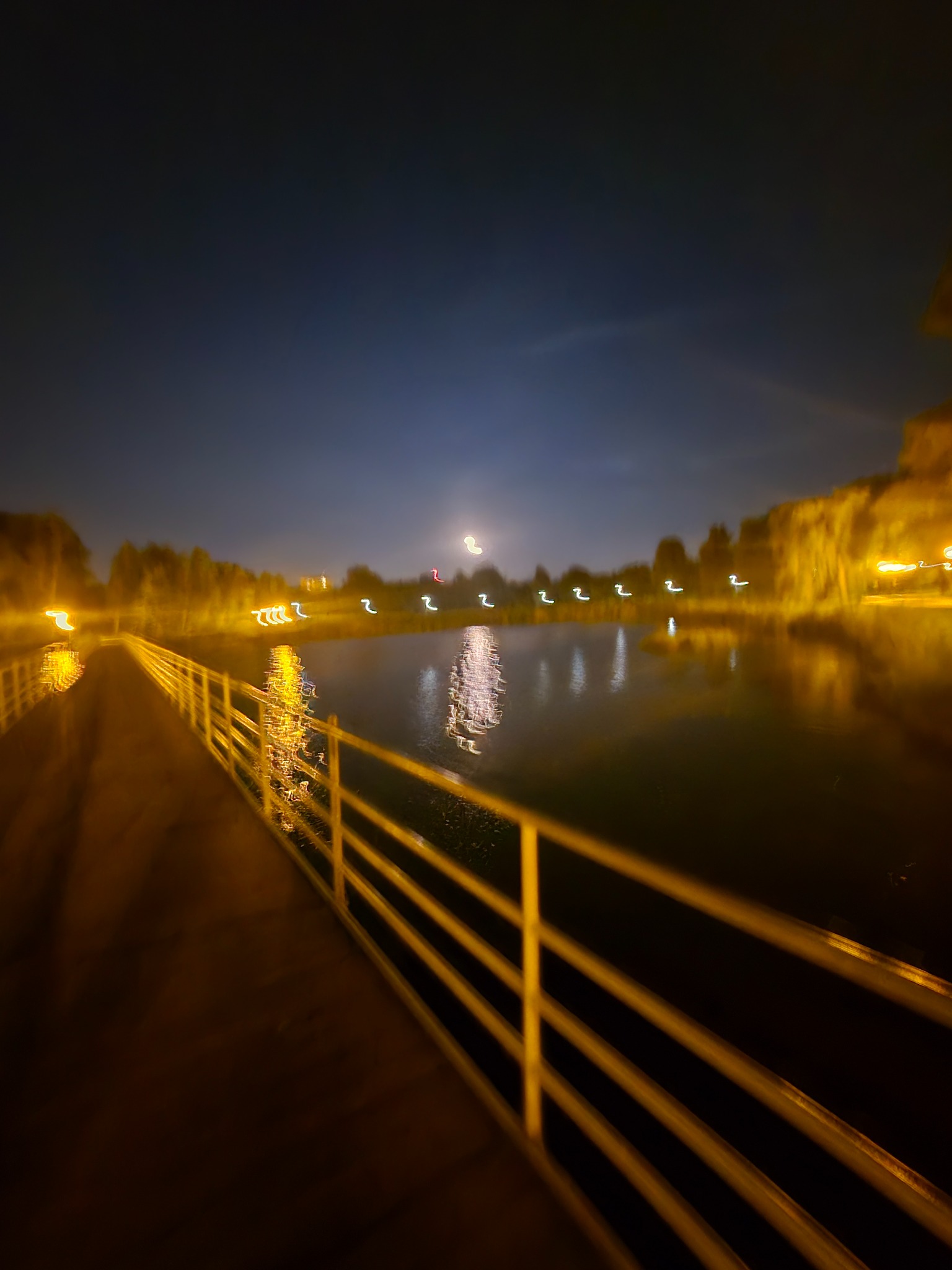 Nocny widok na oświetlony pomost nad jeziorem, z widocznym odbiciem świateł w wodzie. Ujęcie z perspektywy, z żółtą barierką w pierwszym planie.
