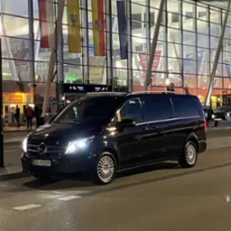 Czarny van Mercedes V-klasy z włączonymi światłami parkuje przed nowoczesnym budynkiem lotniska z flagami. Tablica rejestracyjna widoczna.