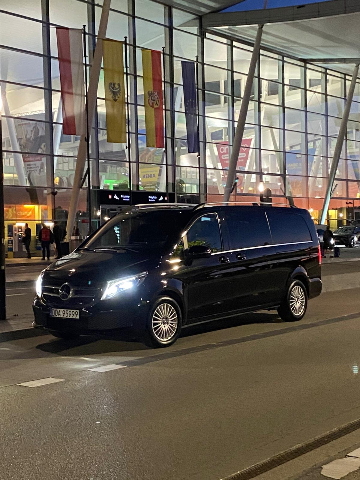 Czarny van Mercedes V-klasy z włączonymi światłami parkuje przed nowoczesnym budynkiem lotniska z flagami. Tablica rejestracyjna widoczna.
