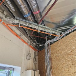 MODENA - Sufit z widocznymi kablami elektrycznymi w peszlach i rurach wentylacyjnych. Kable poprowadzone wzdłuż ściany, z zamontowanymi uchwytami. Widoczny znak ewakuacyjny.