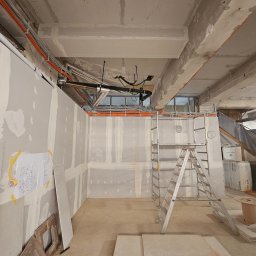 MODENA - Wnętrze w trakcie prac wykończeniowych: ściany z płyt gipsowych, widoczne instalacje pod sufitem, drabina, materiały budowlane. Plan instalacji na ścianie.