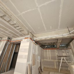 MODENA - Wnętrze w trakcie prac wykończeniowych: widoczne płyty gipsowo-kartonowe na ścianach i suficie, instalacje elektryczne w peszlach oraz drabina.