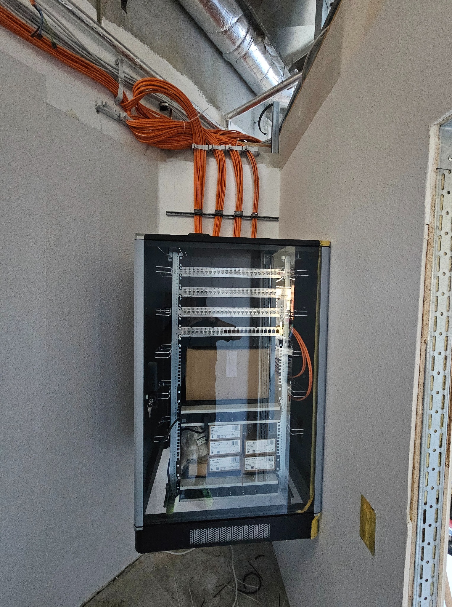 Zamontowana szafa rack z widocznymi kablami pomarańczowymi i elementami instalacji elektrycznej w surowym wnętrzu. Widoczne przewody i elementy montażowe.