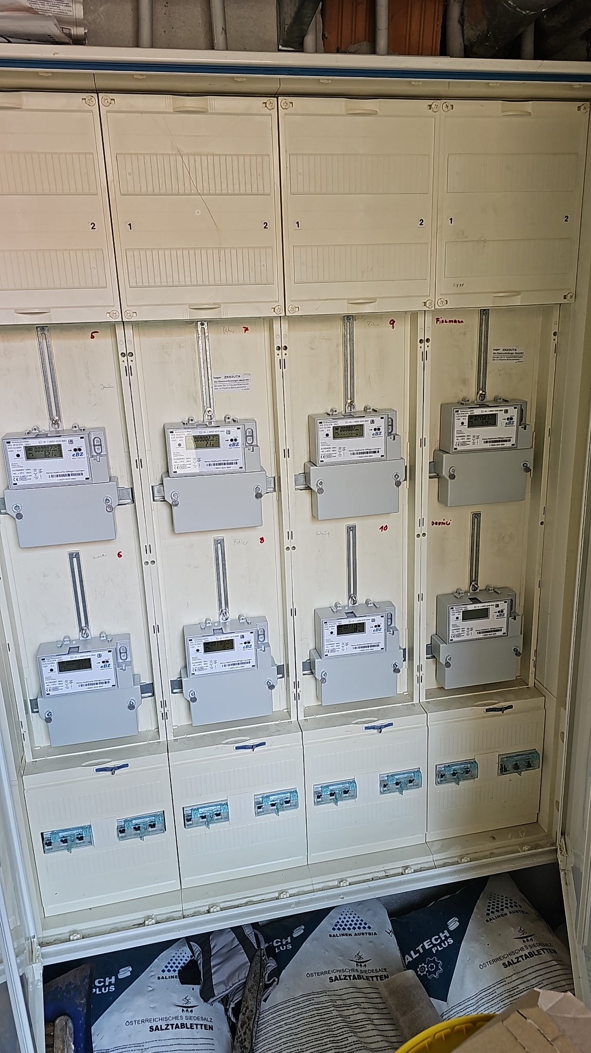 Szafka rozdzielcza z licznikami elektrycznymi w rzędach, widoczne zabezpieczenia i worki z solą tabletkowaną na dole. Ujęcie z bliska, detale instalacji.