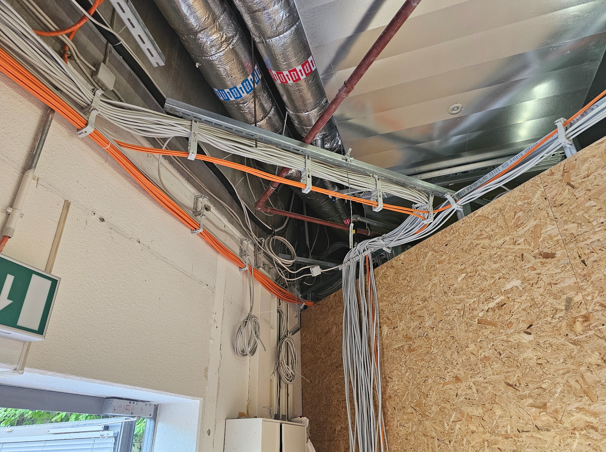 Sufit z widocznymi kablami elektrycznymi w peszlach i rurach wentylacyjnych. Kable poprowadzone wzdłuż ściany, z zamontowanymi uchwytami. Widoczny znak ewakuacyjny.