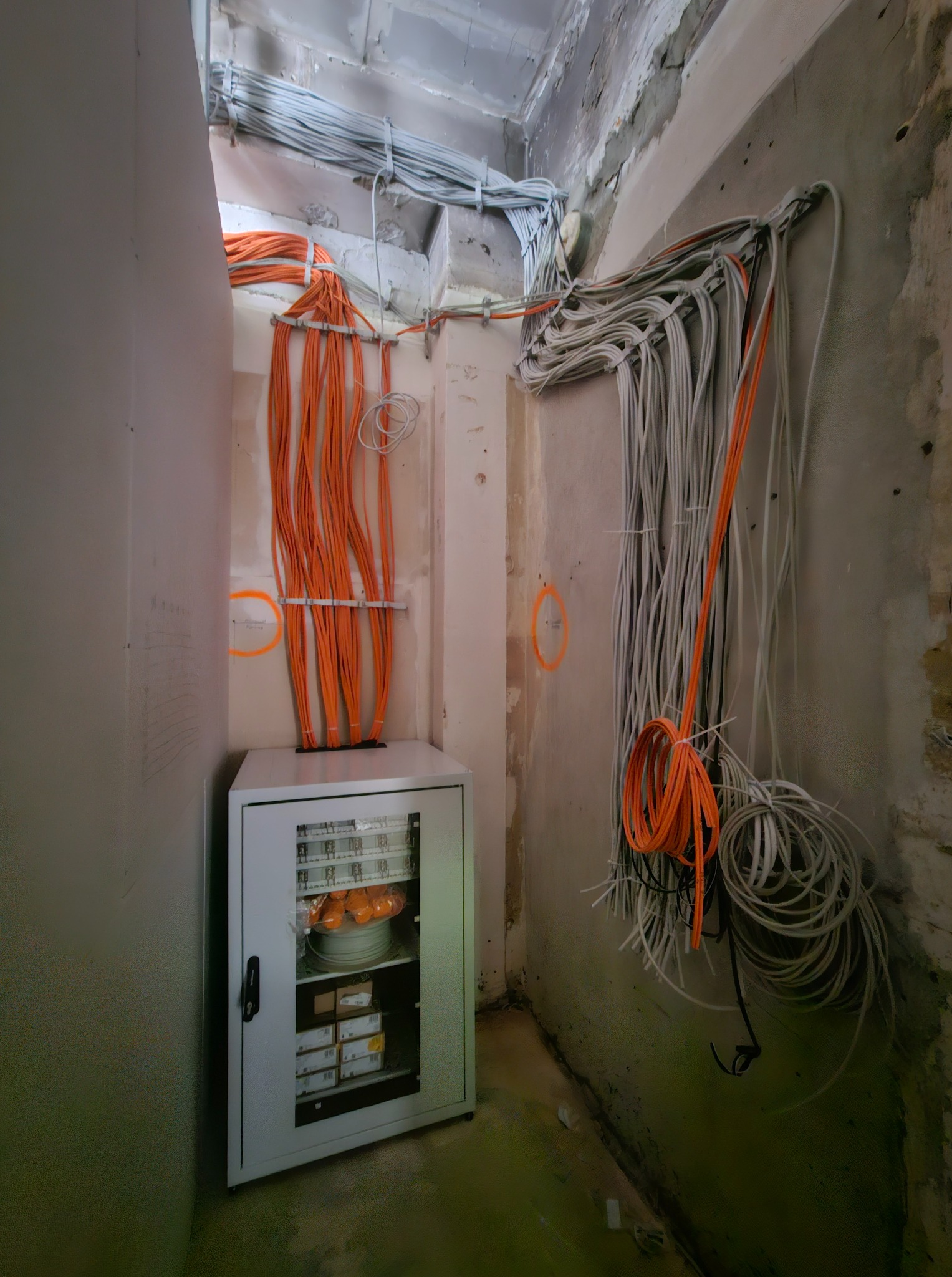 Rozprowadzenie kabli elektrycznych w surowym wnętrzu, widoczna szafka z elementami instalacji. Pomarańczowe i szare przewody prowadzone wzdłuż ścian i sufitu.