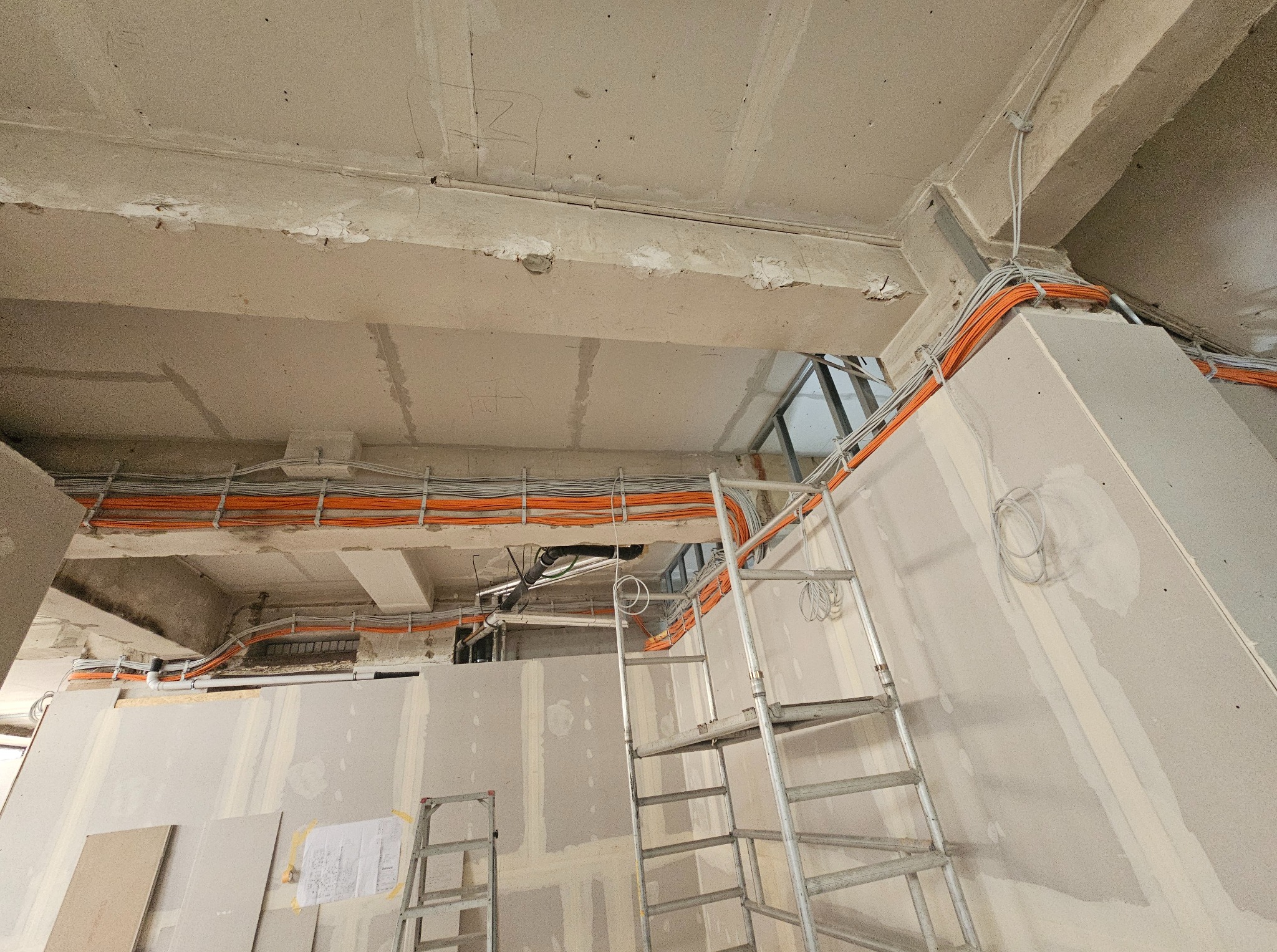 Wnętrze w trakcie prac wykończeniowych: widoczne kable elektryczne ułożone pod sufitem, ściany przygotowane do malowania, drabiny. Surowy stan.