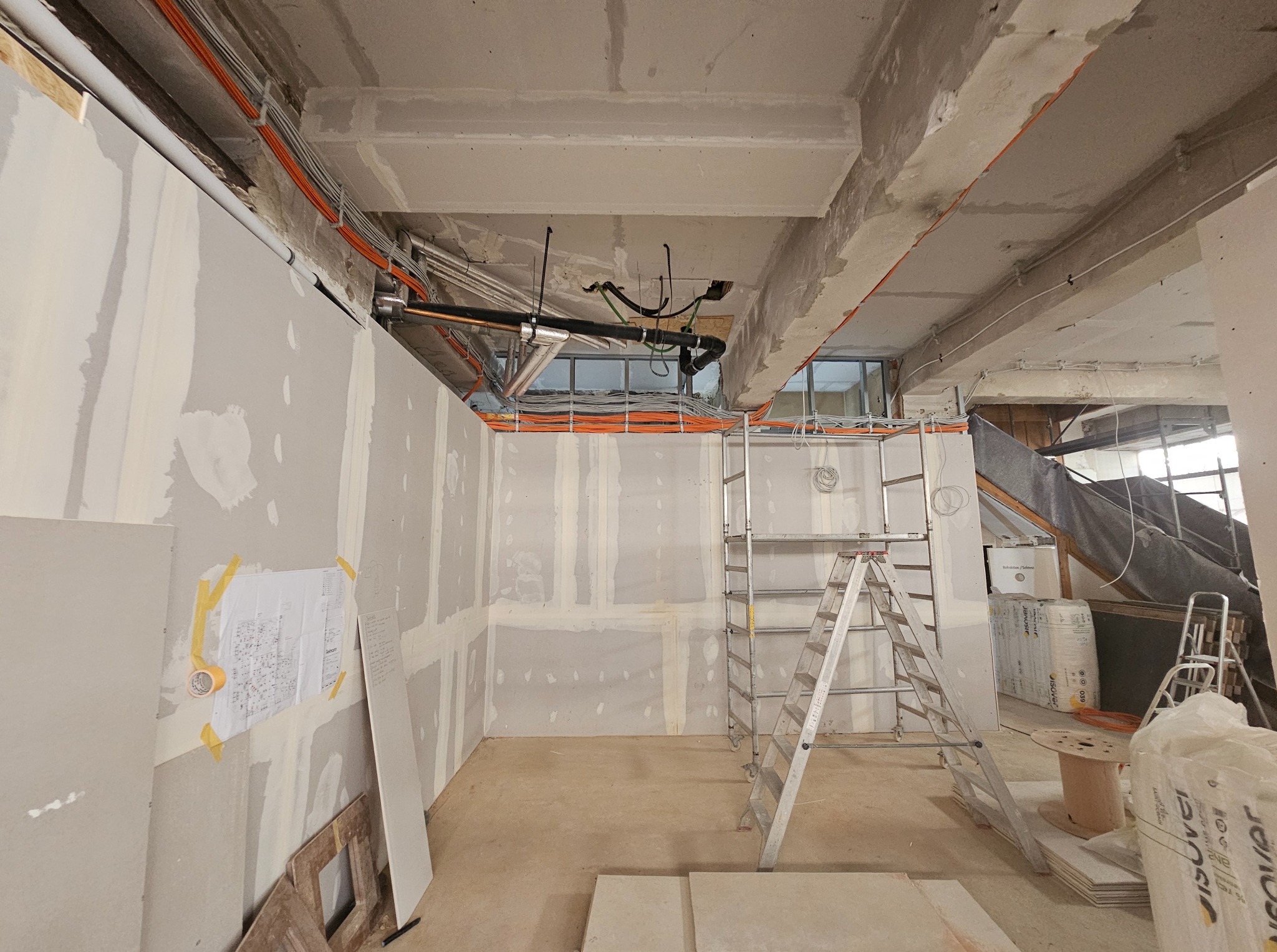 Wnętrze w trakcie prac wykończeniowych: ściany z płyt gipsowych, widoczne instalacje pod sufitem, drabina, materiały budowlane. Plan instalacji na ścianie.