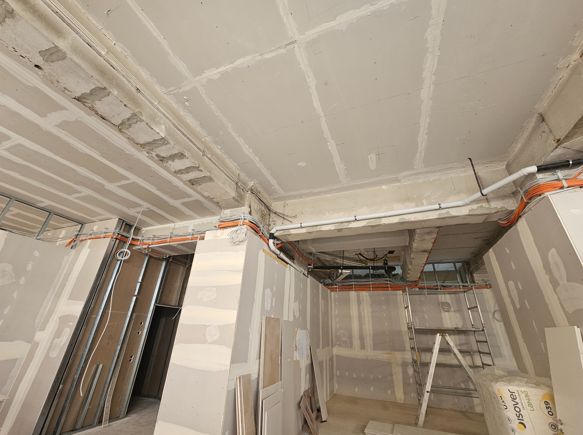 Wnętrze w trakcie prac wykończeniowych: widoczne płyty gipsowo-kartonowe na ścianach i suficie, instalacje elektryczne w peszlach oraz drabina.