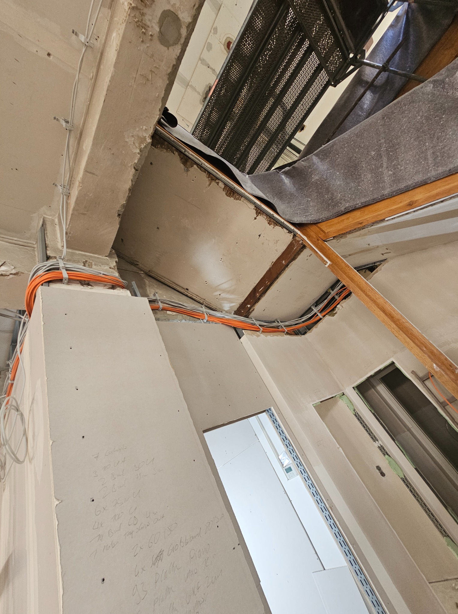 Instalacja elektryczna: Pomarańczowe kable prowadzone wzdłuż sufitu i ścian w trakcie remontu. Widoczne płyty kartonowo-gipsowe i elementy konstrukcyjne.