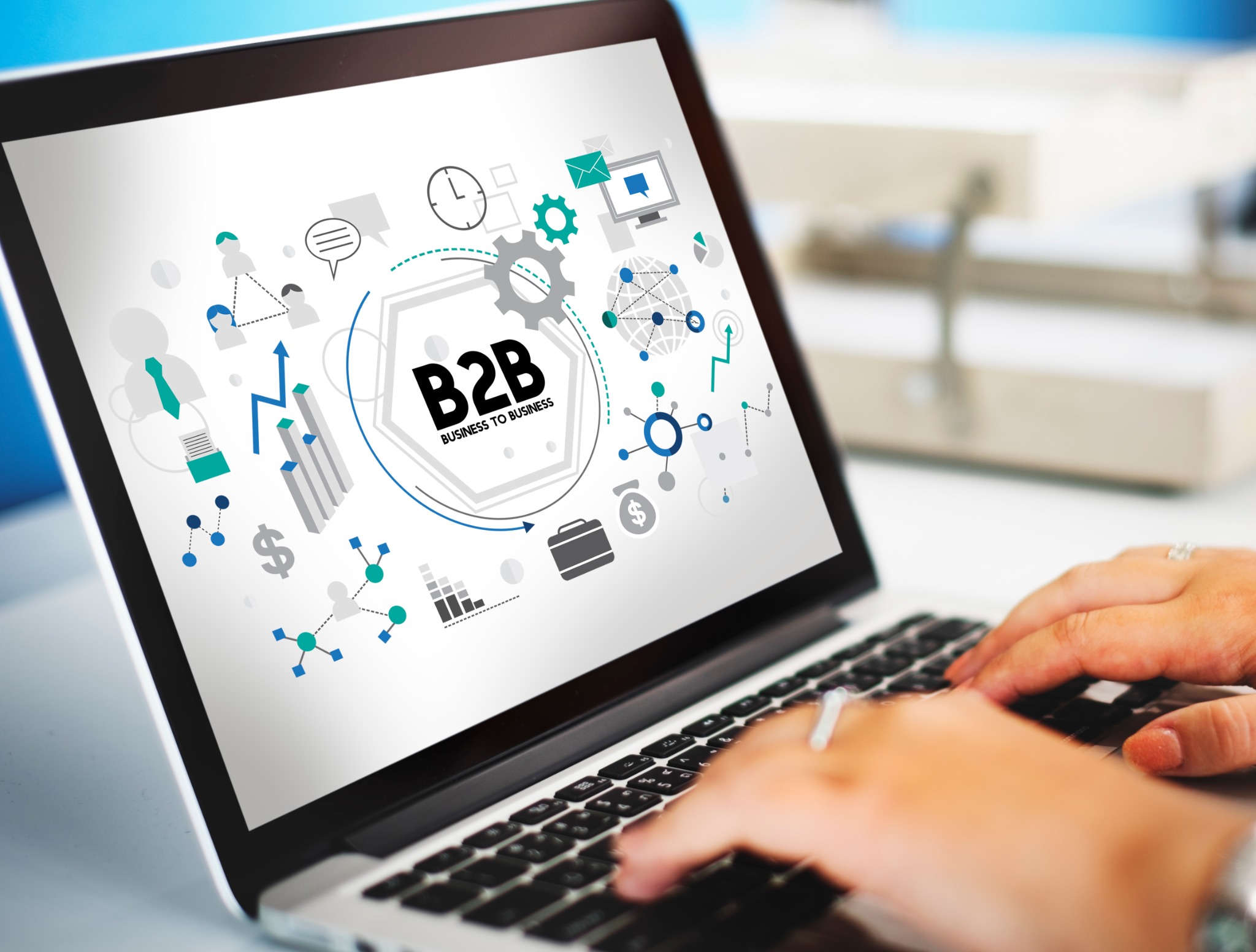 Laptop z grafiką B2B i dłońmi na klawiaturze, sugerujący moduł Business-to-Business. Widoczne ikony: zegar, koło zębate, wykresy, sieć powiązań i torba z dolarem.