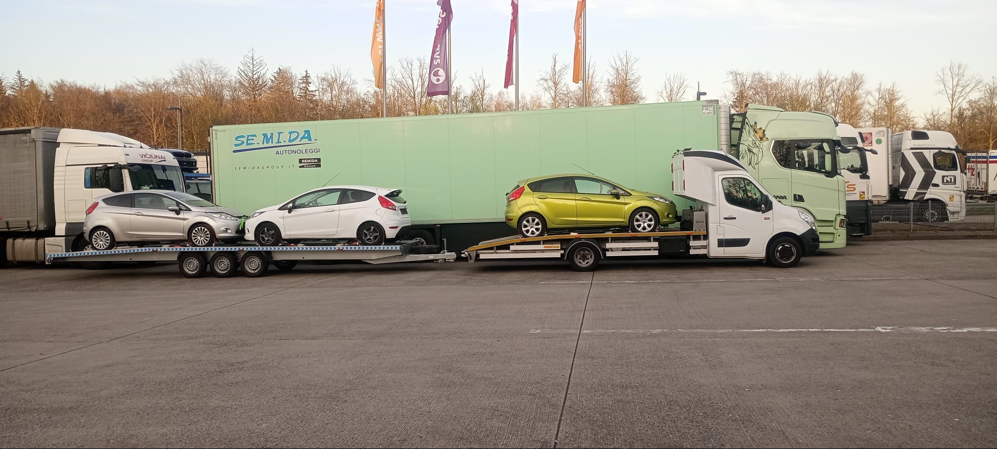 Laweta przewozi trzy samochody Ford Fiesta w kolorach: srebrny, biały i limonkowy. Na naczepie widoczne logo firmy transportowej. W tle drzewa i inne ciężarówki.