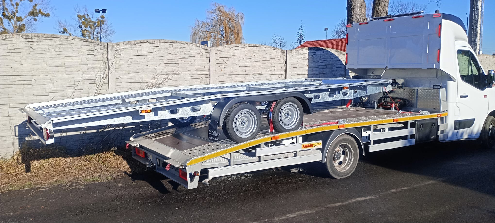 Laweta: biały samochód z dwupoziomową przyczepą do transportu aut, widok z boku na tle betonowego muru i drzew bez liści, w słoneczny dzień.