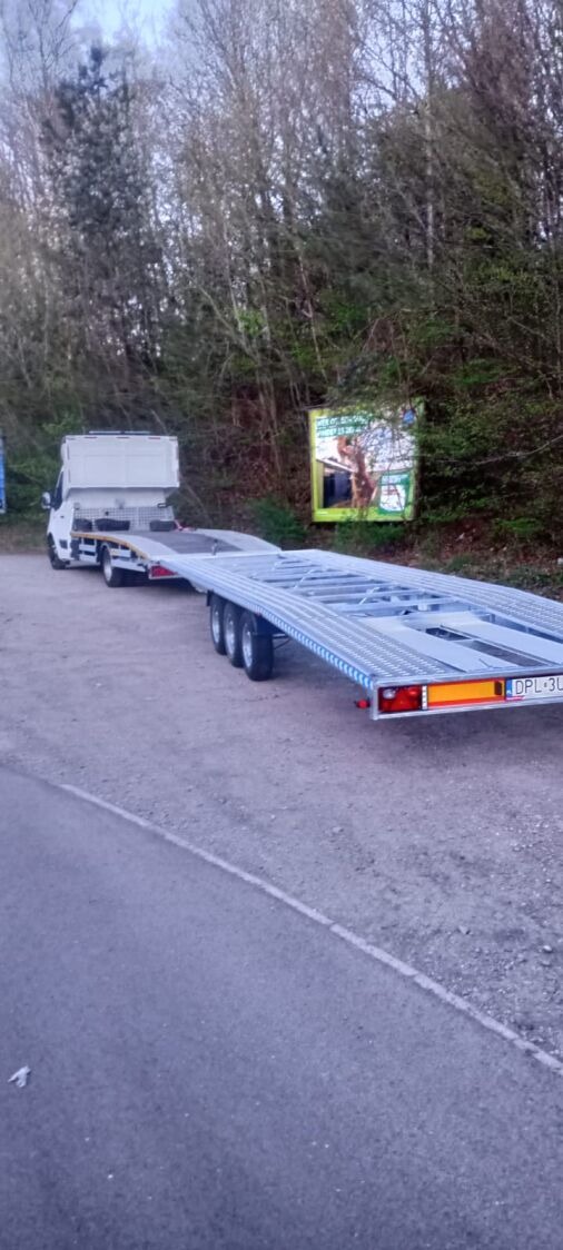 Laweta z platformą na trzy auta, zaparkowana przy drodze, na tle drzew i billboardu. Widoczny numer rejestracyjny DPL 3U.