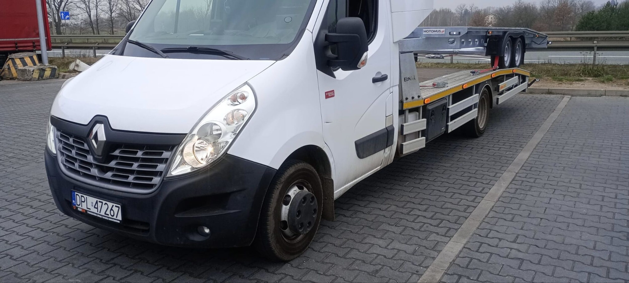 Biała laweta Renault Master z platformą MOTOMUS.pl na parkingu, widok z przodu i boku. Tablica rejestracyjna DPL 47267. Widoczne drzewa w tle.