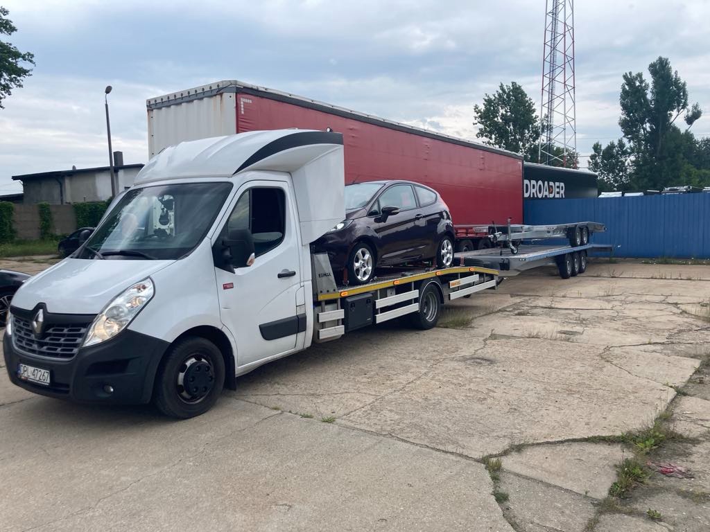 Biała laweta Renault z fioletowym autem na platformie i przyczepą na betonowym placu z trawą, w tle czerwona naczepa i niebieskie ogrodzenie.