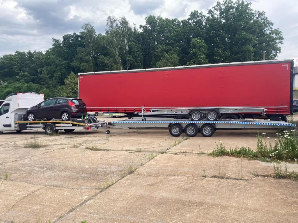 Laweta z brązowym autem osobowym na platformie, zaparkowana obok czerwonej naczepy na betonowym placu, w tle drzewa i pochmurne niebo.