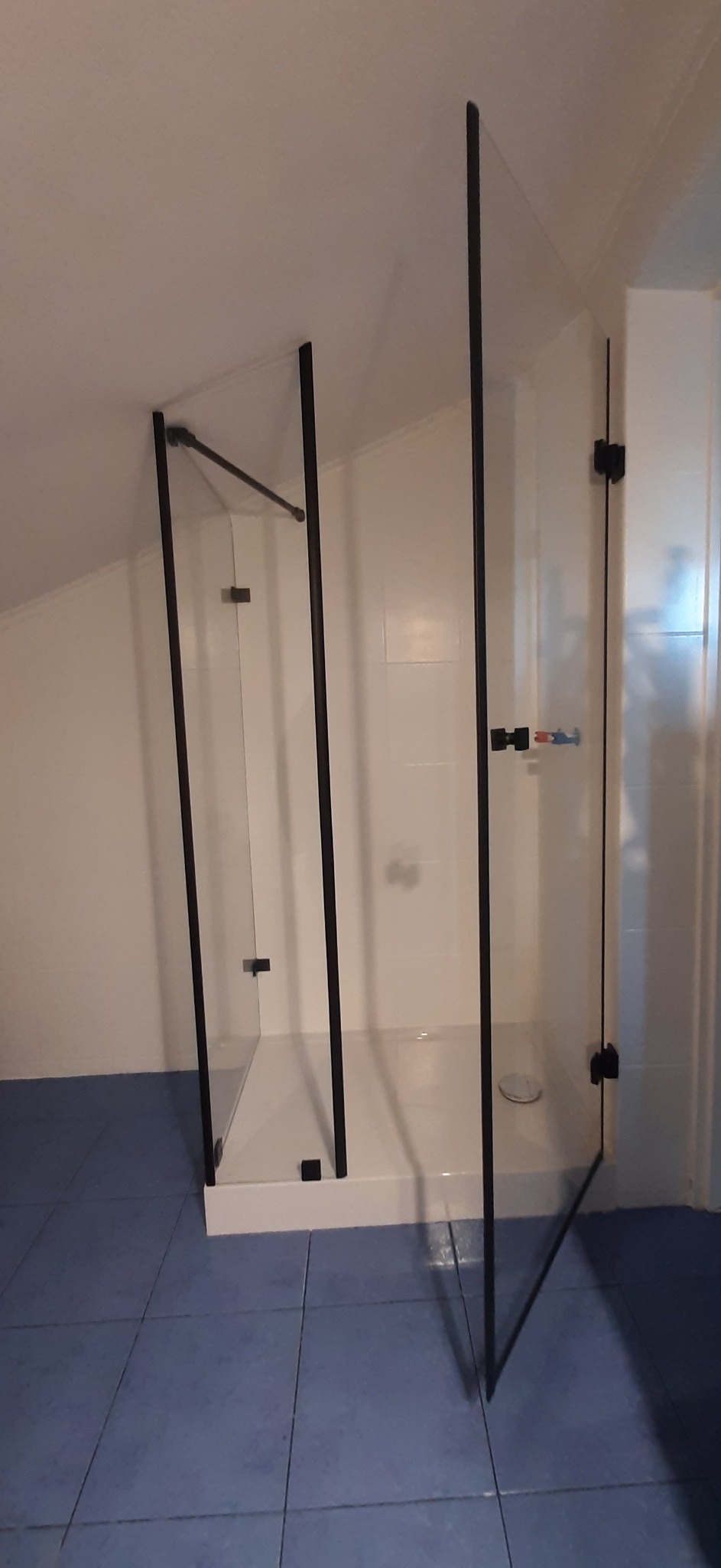 Kabina prysznicowa ze szklanymi drzwiami i czarnymi profilami w łazience na poddaszu. Widoczny biały brodzik i niebieska podłoga.