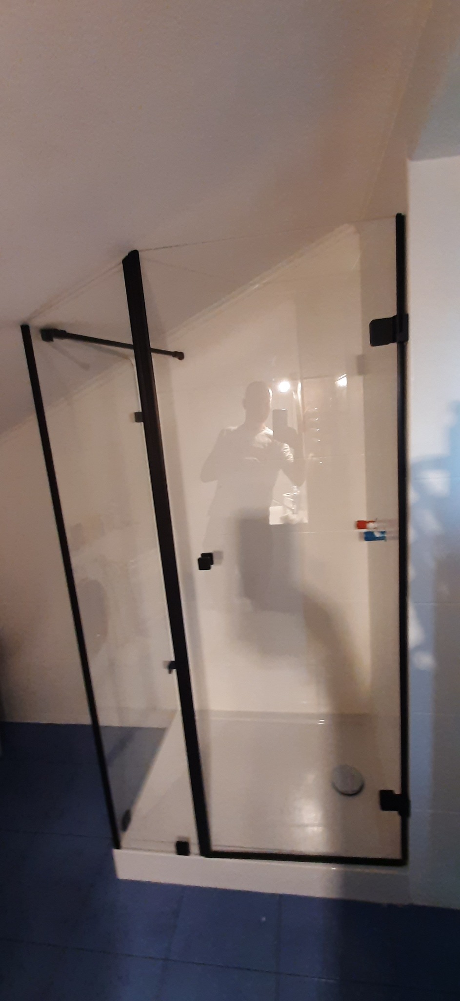 Kabina prysznicowa ze szkła z czarnymi profilami, zamontowana w łazience na poddaszu ze skośnym sufitem. Widoczne detale wykończenia i odbicie osoby robiącej zdjęcie.