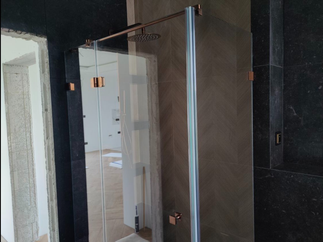 Nowoczesna kabina prysznicowa z transparentnego szkła, z metalowymi okuciami w kolorze miedzi, na tle płytek w odcieniach szarości i czerni. Widoczny fragment łazienki w trakcie wykończenia.