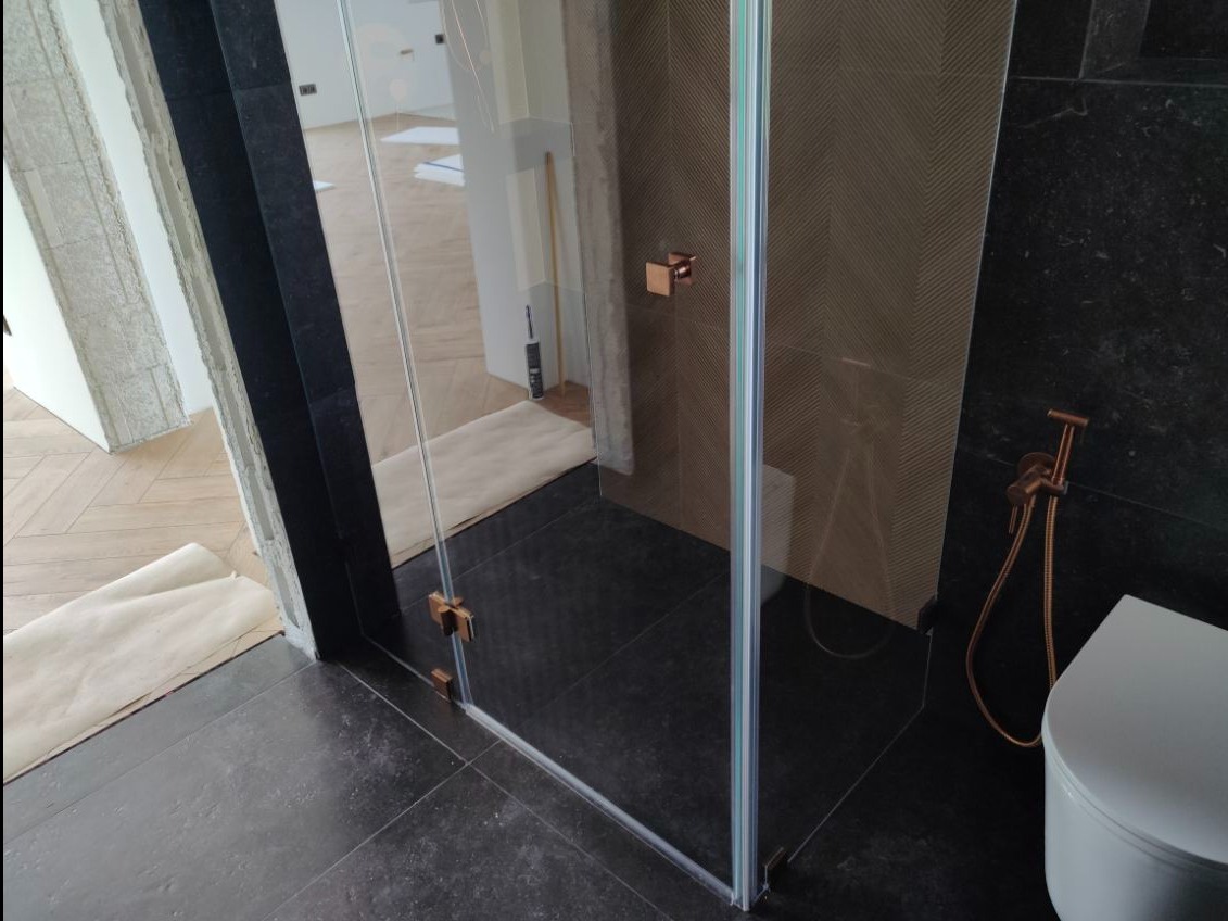 Nowoczesna kabina prysznicowa z przezroczystego szkła, z okuciami w kolorze miedzi, na tle ciemnych płytek w łazience. Widoczne elementy wykończenia wnętrza.