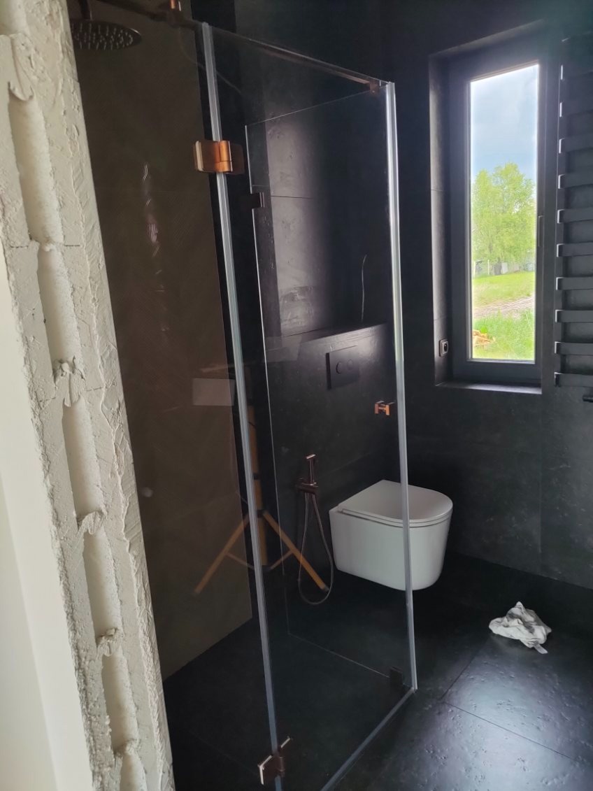 Nowoczesna kabina prysznicowa ze szkła z miedzianymi okuciami w łazience z ciemnymi płytkami i oknem. Widoczny fragment surowej ściany.