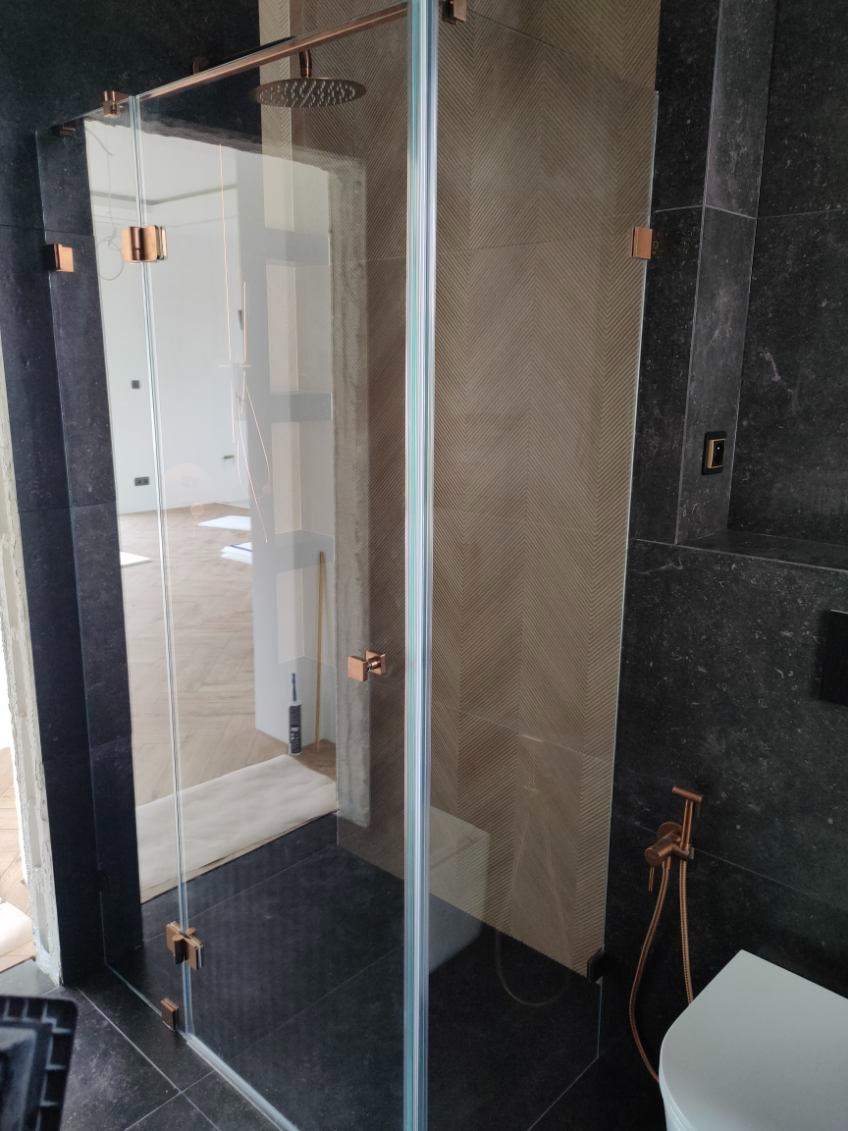 Nowoczesna kabina prysznicowa ze szkła hartowanego, z okuciami w kolorze miedzi, na tle płytek w geometryczny wzór. Widoczny fragment toalety i deszczownica.