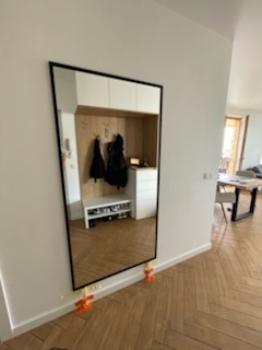 Duże, prostokątne lustro w czarnej ramie, oparte o ścianę w przedpokoju z widoczną zabudową meblową i parkietem w jodełkę. Pomarańczowe podkładki zabezpieczają dół lustra.