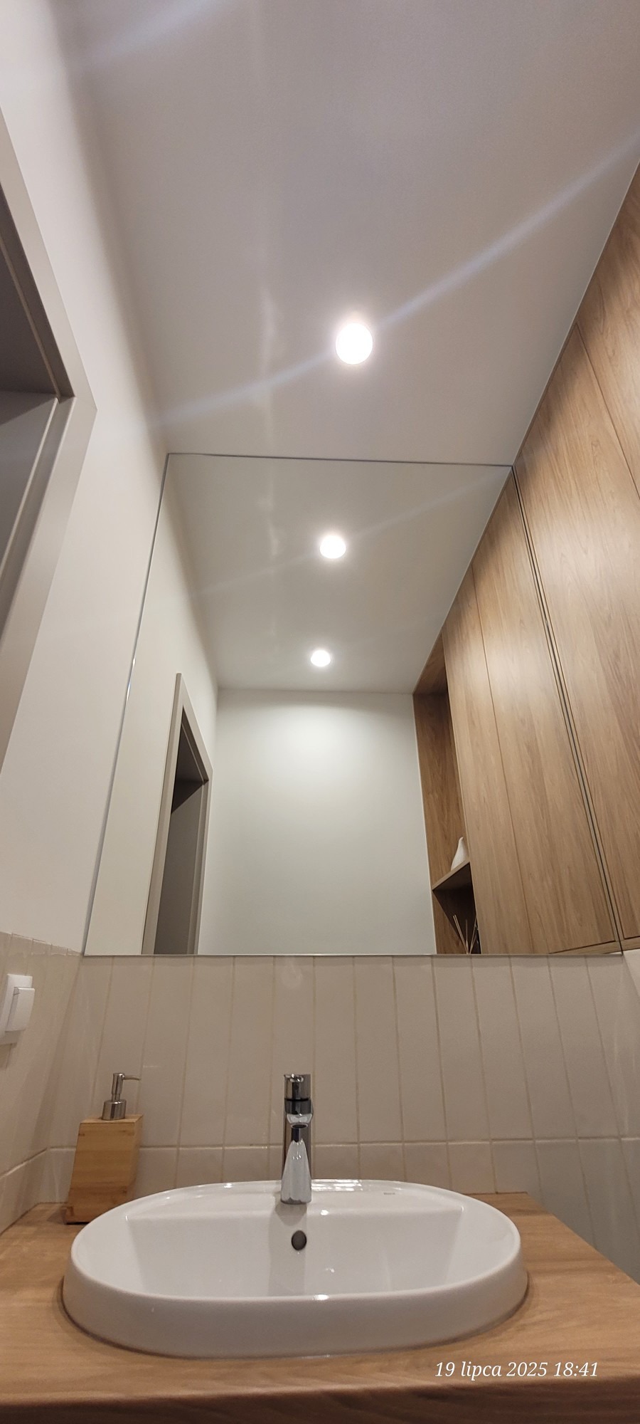 Nowoczesna łazienka z dużym, prostokątnym lustrem nad umywalką, odbijającym wnętrze z oświetleniem punktowym i drewnianą zabudową. Minimalistyczny design.