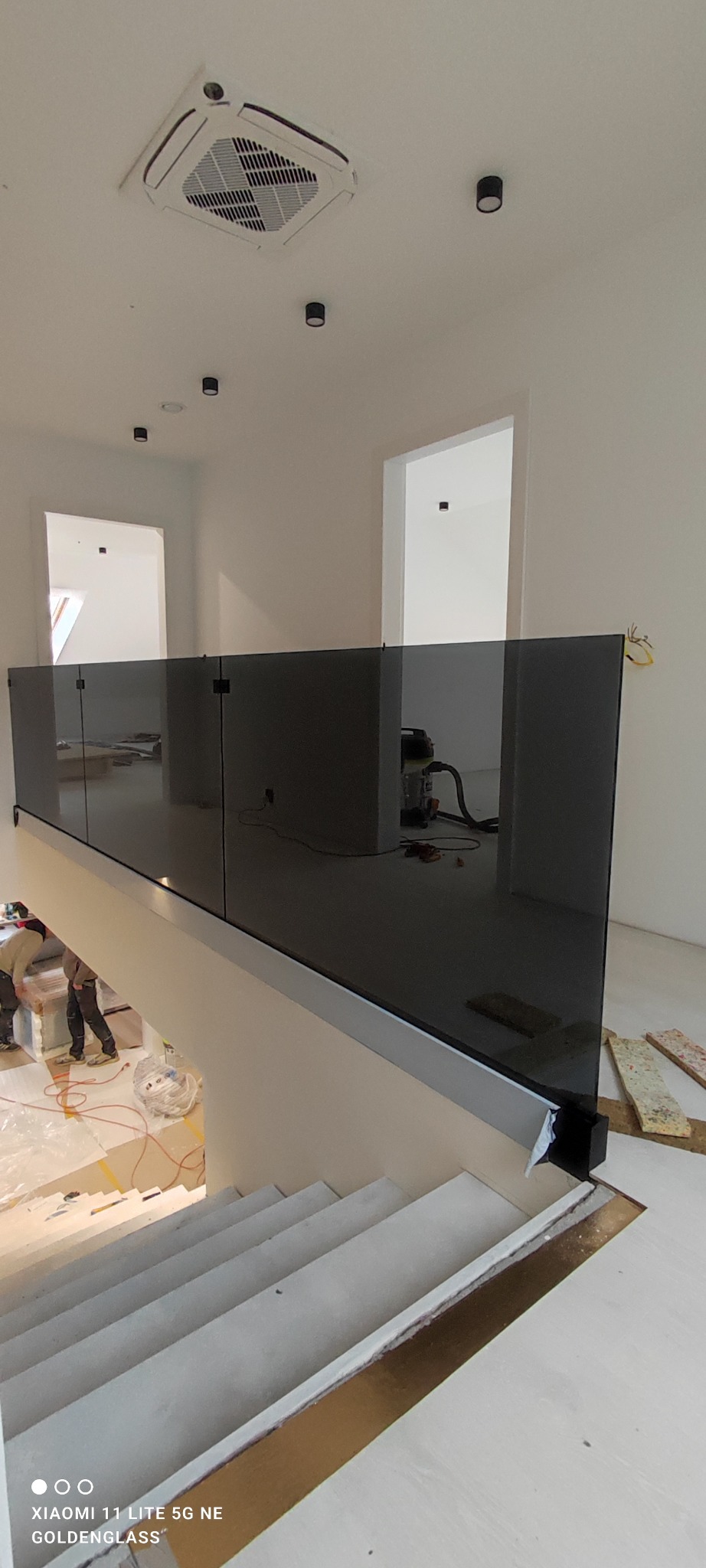 Nowoczesna, przyciemniana balustrada szklana na schodach w jasnym wnętrzu. Widoczne elementy montażowe i częściowo ekipa montażowa. Minimalistyczny design.