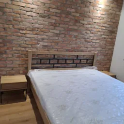 Drewniane łóżko z szafkami nocnymi na tle ściany z czerwonej cegły, oświetlone punktowo. Materac w folii ochronnej. Widok z przodu, z bliska.