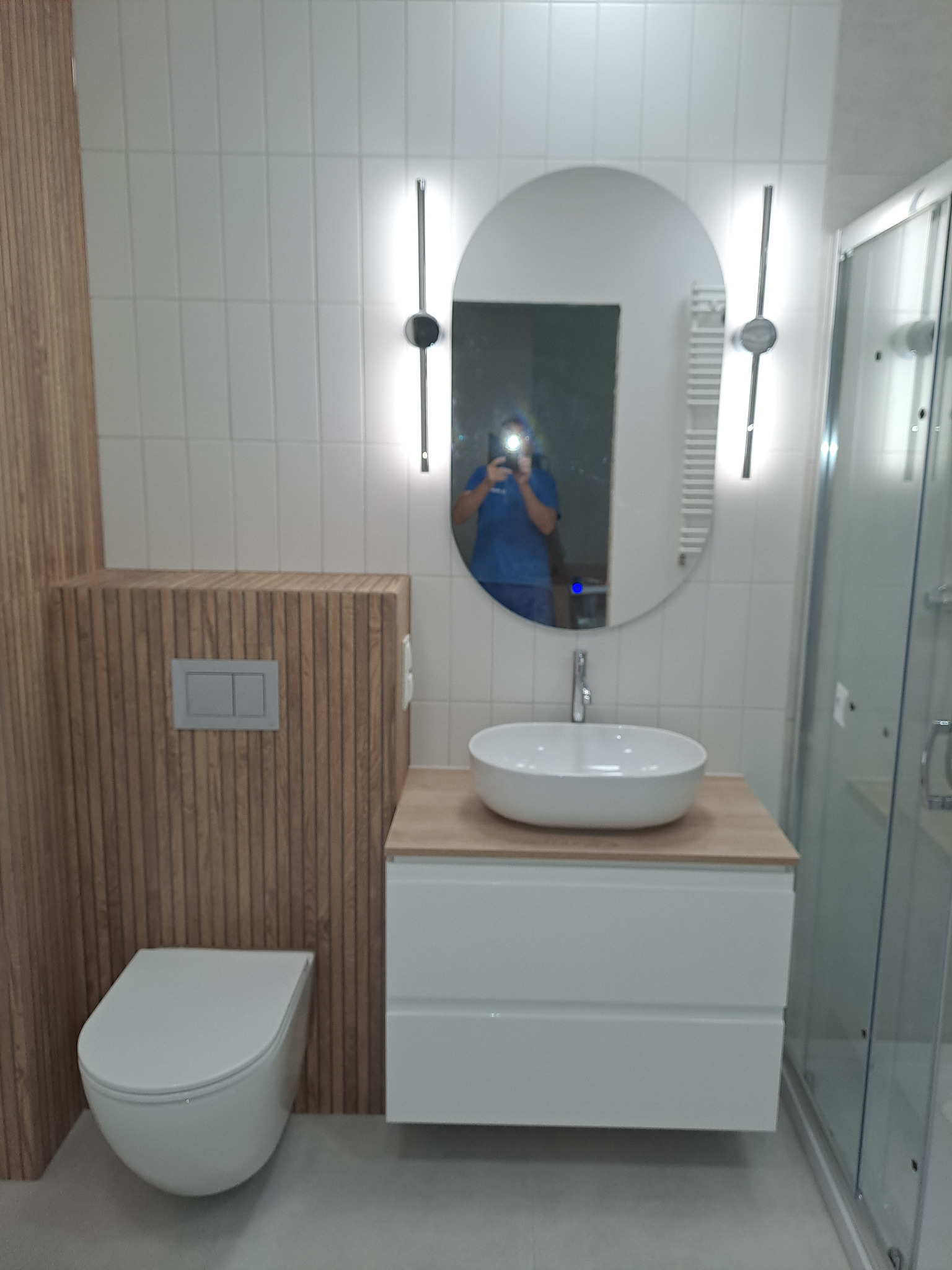 Nowoczesna łazienka z owalnym lustrem podświetlanym LED, białą umywalką nablatową i zabudową WC z drewnianymi lamelami. Kabina prysznicowa ze szklanymi drzwiami.