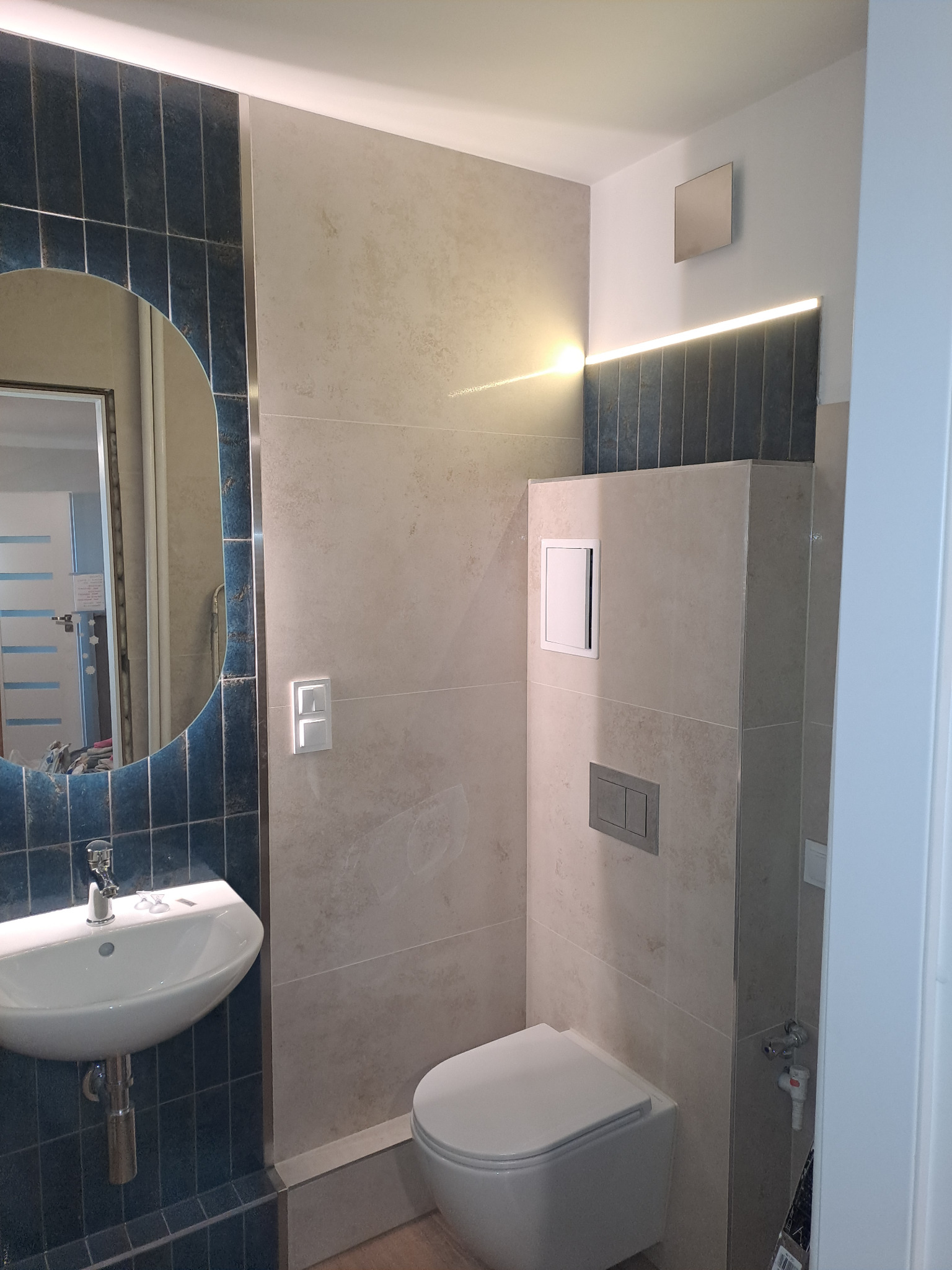 Wykończona łazienka z umywalką, toaletą i podświetleniem LED. Ciemne i jasne płytki tworzą kontrast. Lustro o owalnym kształcie.