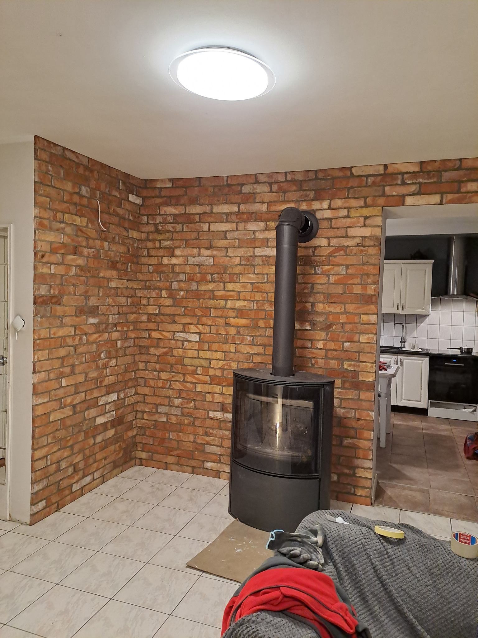 Ceglana ściana z kominkiem typu koza w rogu pokoju, widok na kuchnię w tle. Oświetlenie sufitowe. Częściowo widoczny narożnik sofy z akcesoriami.