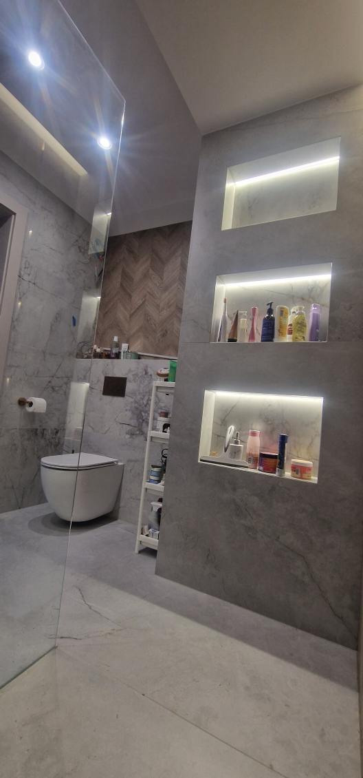 Nowoczesna łazienka z marmurowymi płytkami, oświetlonymi wnękami na kosmetyki i toaletą. Widoczna szklana ścianka prysznicowa i drewniana ściana w jodełkę w tle.