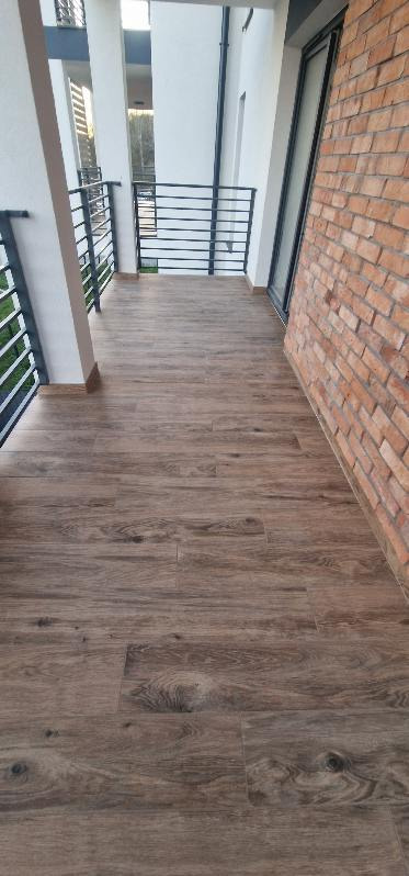 Balkon z podłogą z płytek imitujących drewno i ceglaną ścianą. Czarne metalowe barierki. Ujęcie z góry, widok na zewnątrz. Minimalistyczny styl.