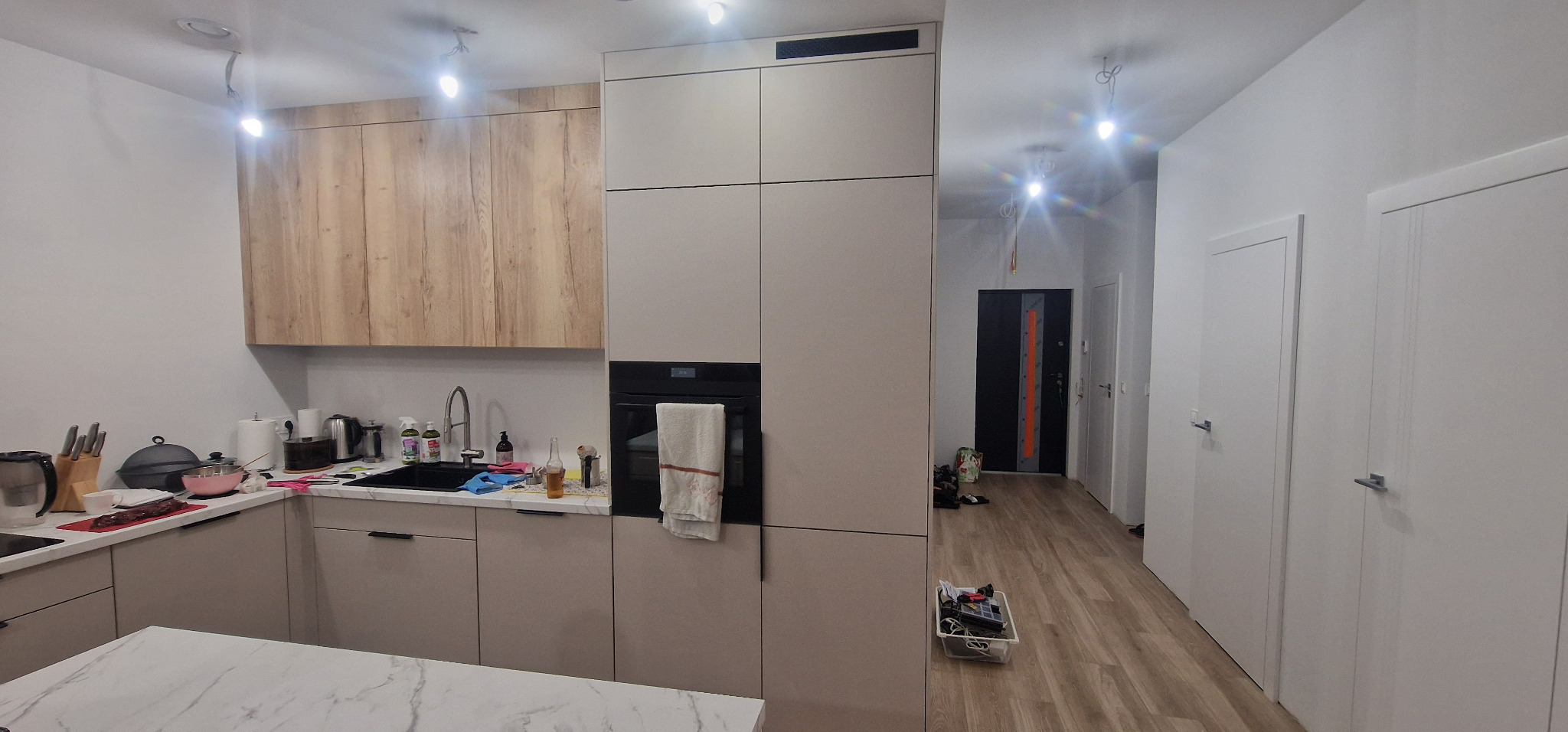 Nowoczesna kuchnia z jasnymi szafkami, drewnianymi akcentami i czarnym piekarnikiem. Widok na korytarz z białymi drzwiami i podłogą imitującą drewno.