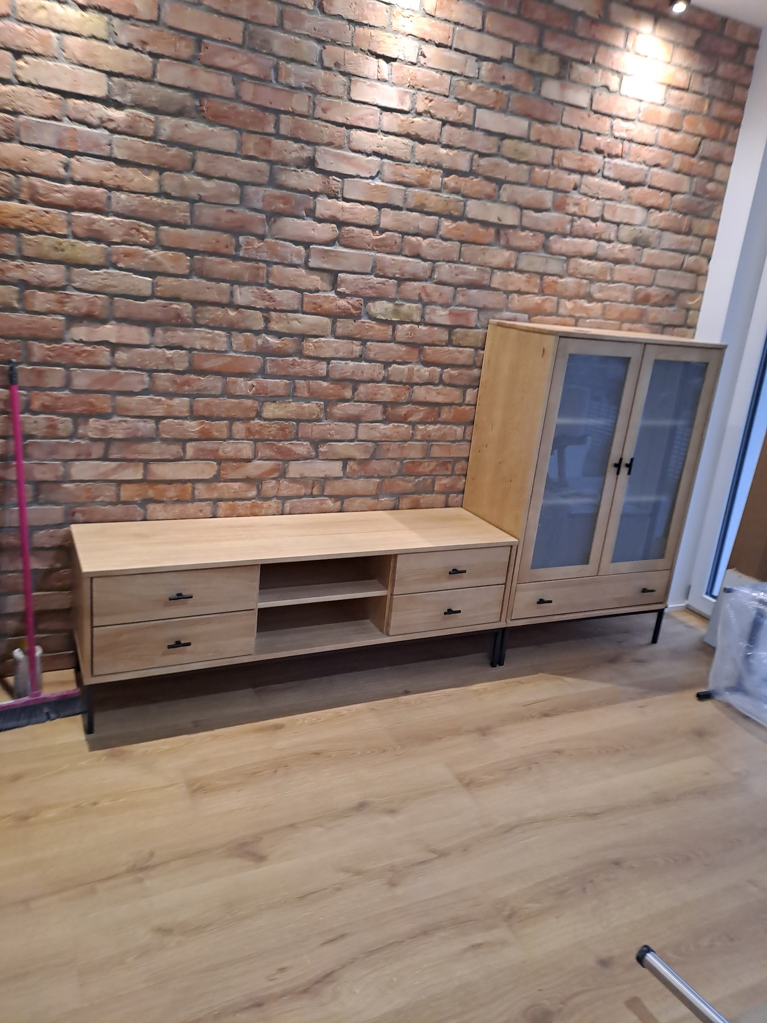 Jasne, drewniane meble (szafka RTV i witryna) stoją na podłodze z paneli na tle ściany z czerwonej cegły, tworząc nowoczesne wnętrze.