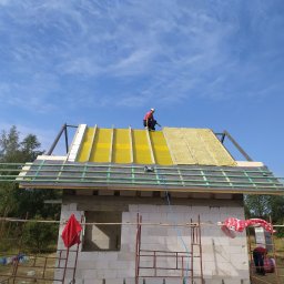Usługi budowlane i ogrodnicze - Montaż dachu na domu w budowie: dekarz na żółtej membranie i izolacji z wełny mineralnej, widoczne rusztowania i narzędzia dekarskie, błękitne niebo w tle.