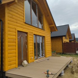 Usługi budowlane i ogrodnicze - Żółty dom z drewnianą elewacją i czarnym dachem. Widoczny taras w trakcie budowy, narzędzia i materiały budowlane. Ujęcie z boku, w tle kolejne domy.